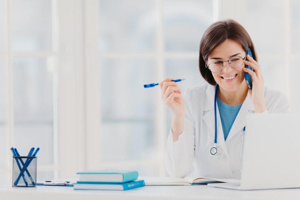 Imagem de uma médica sentada em frente a mesa do seu consultório, falando ao celular e lendo informações. 